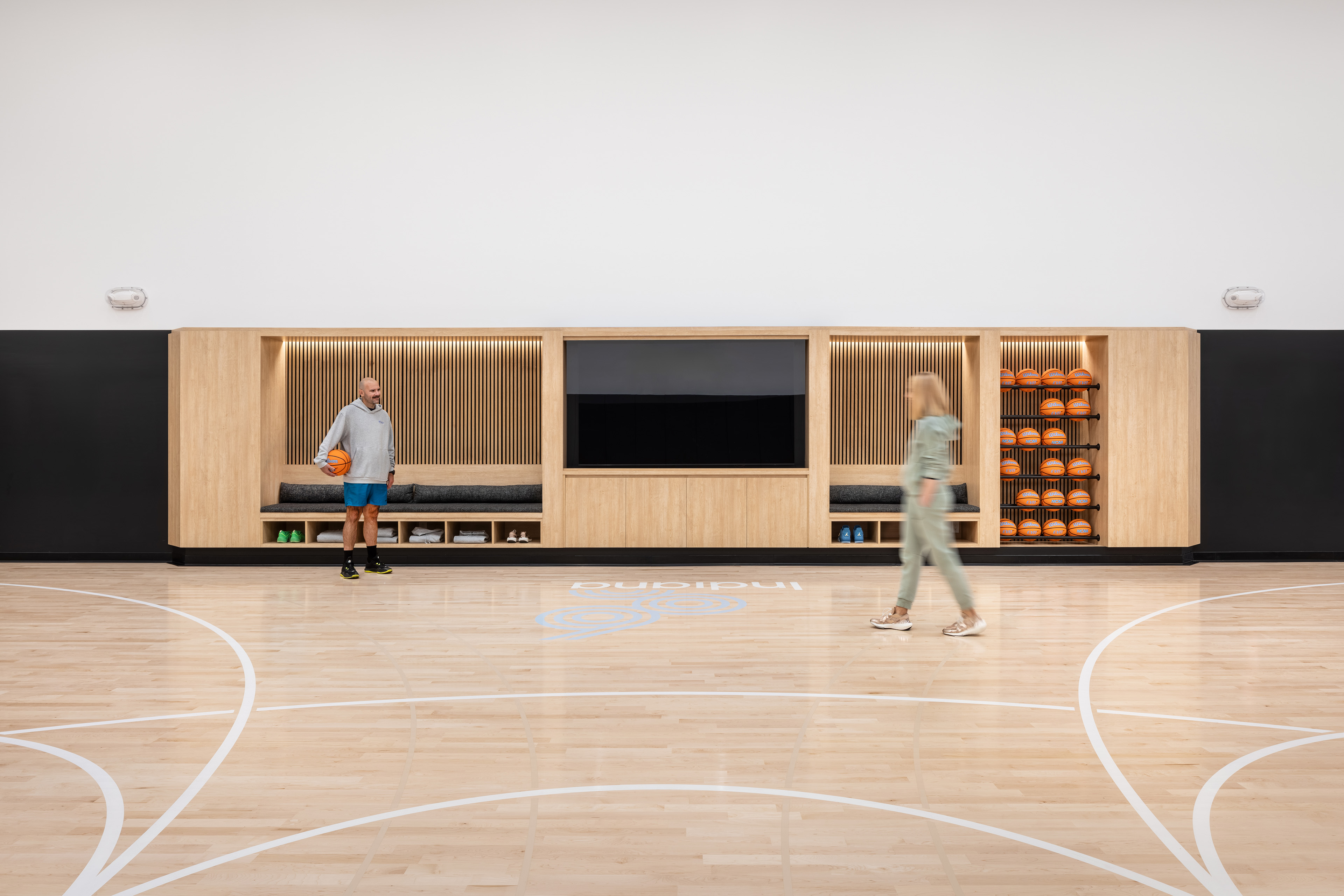 A straight-on shot of a large, custom light-wood media and storage unit built into a basketball court's wall. A man holding a basketball stands talking to a woman who is walking past. The unit features built-in bench seating with slatted wood backdrops, a large center cutout for a flat-screen TV, and an open rack filled with rows of basketballs on the right.