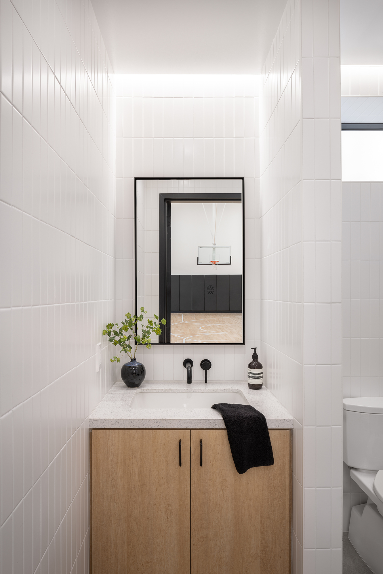 A minimalist restroom vanity alcove featuring white vertically tiled walls and a light wood cabinet. A black-framed rectangular mirror sits above a white quartz countertop, reflecting an open doorway that looks out onto the basketball court. The sink has matte black wall-mounted faucets, a black towel, a soap dispenser, and a small black vase with green foliage.
